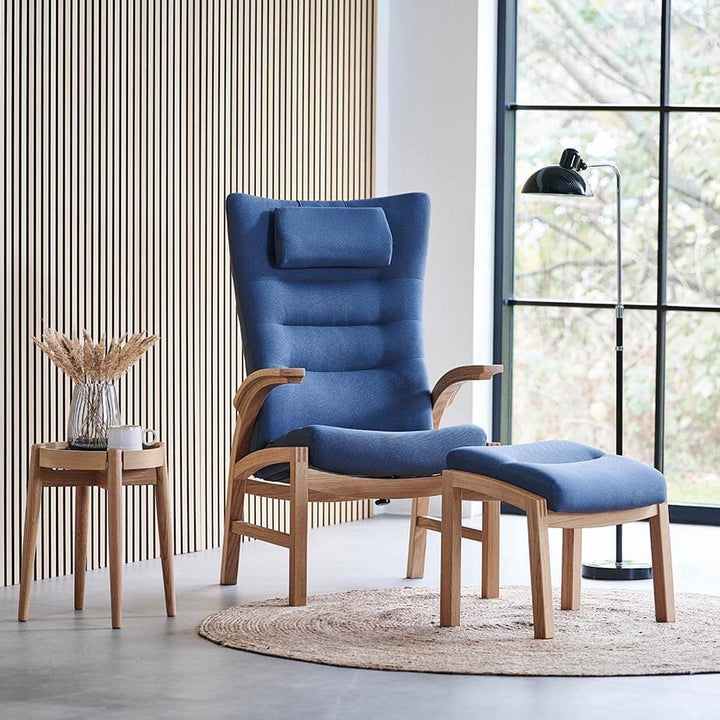 Lænestol med skammel, model Cantate 6011, med stel i natur eg og polster i bæredygtigt støvet blåt tekstil, stående foran stort vinduesparti og væg med egetræslameller
